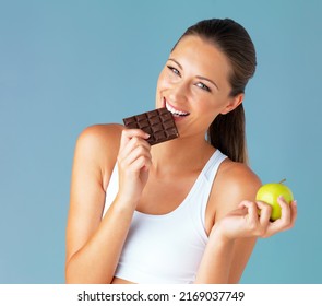 My Diet Makes Room For The Occasional Indulgence. Studio Shot Of A Fit Young Woman Holding An Apple While Taking A Bite Of Chocolate Against A Blue Background.