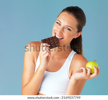 My diet makes room for the occasional indulgence. Studio shot of a fit young woman holding an apple while taking a bite of chocolate against a blue background.