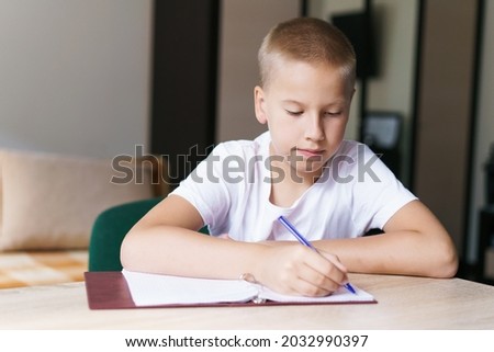 My favorite subject. Handsome smiling blonde boy is working and writing in his notebook while sitting at table, doing his homework on the school subject. Caucasian boy writes in a notebook with a pen
