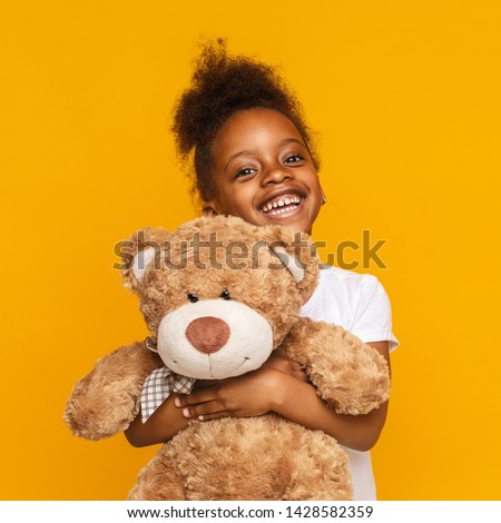 My favorite toy. Cute little african girl hugging her teddy bear and smiling, orange studio background