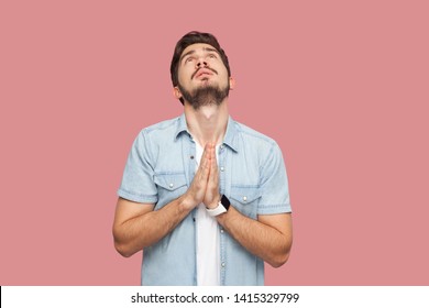 My God please help me. Portrait of hopeful sad handsome bearded young man in blue casual style shirt standing looking up and praying with worry face. indoor studio shot, isolated on pink background.