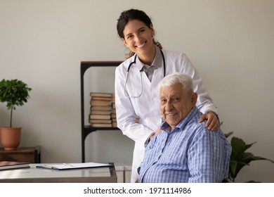 My Health Is In Safe Hands. Happy Young Latin Woman Gp Hug Shoulders Of Old Man Visitor Glad To Help Him Feel Well. Smiling Doc And Retired Patient Look At Camera Posing For Portrait In Doctor Office