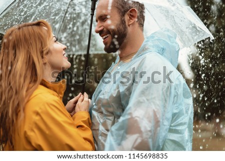 My love. Profile of delighted male and female facing each other under one umbrella. They are enjoying rain together with smile and content