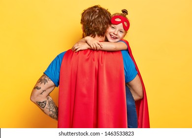 My lovely super dad. Affectionate small child cuddles father with love, wears red mask, dressed in superhero costumes, confident in defense and dads strength, play together, isolated on yellow wall