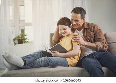 My loving son. Cheerful boy is reading book with interest and smiling. His father is embracing him and stroking his head with fondness - Powered by Shutterstock