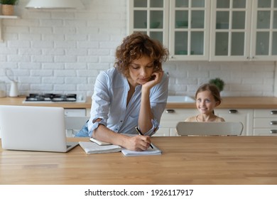 My mommy works from home. Busy mother sit at kitchen table by pc speak on phone take notes while little daughter play near. Female freelancer engaged in multitasking job unable to spend time with kid