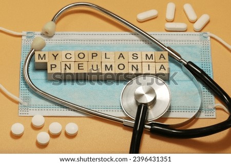 Mycoplasma pneumonia, caused by the bacterium Mycoplasma pneumoniae. The words are laid out with wooden cubes on a surgical face mask. Various pills and a stethoscope are lying around.