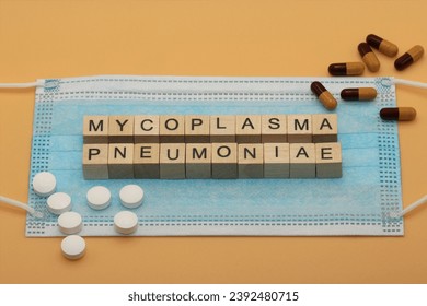 Mycoplasma pneumoniae. It is a human pathogen that causes the disease mycoplasma pneumonia. The words are laid out with wooden cubes on a surgical face mask. There are various pills lying around.