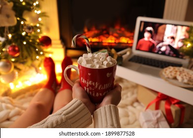 MYKOLAIV, UKRAINE - DECEMBER 23, 2020: Woman with sweet drink watching Harry Potter and Philosopher's Stone movie on laptop near fireplace at home, closeup. Cozy winter holidays atmosphere