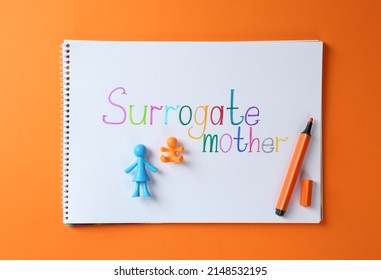 MYKOLAIV, UKRAINE - JANUARY 04, 2022: Notebook With Words Surrogate Mother, Marker And Human Figures On Orange Background, Top View