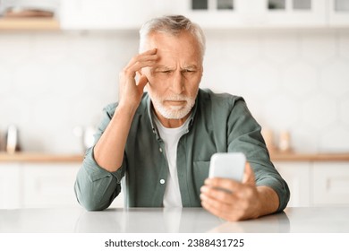 Myopia Concept. Senior man squinting while looking at smartphone screen, elderly gentleman holding mobile phone and trying to read message while sitting at desk in kitchen interior, closeup - Powered by Shutterstock