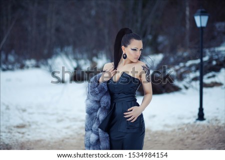 Mysterious art portrait of beautiful brunette woman in fashionable long leather dress and fur coat with fashion hairstyle and make up against the background of winter park. Girl with dragon tattoo

