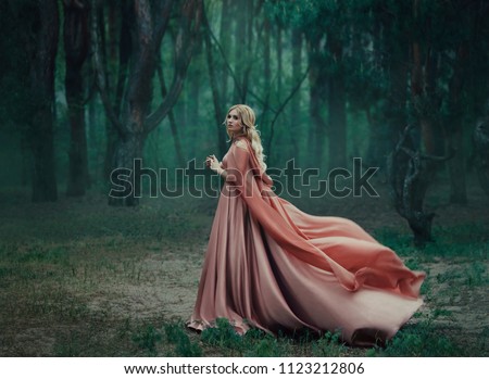 A mysterious blonde girl in a long pink dress with a train and a raincoat that flutters in the wind. The wizard leaves in a forest covered with fog. A background of trees with a haze away. Art photo