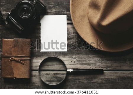 Mysterious detective game concept notebook leather cover sheet white paper felt brown hat portable mirrorless microthird digital photo camera big vintage magnifier isolated on black aged wood table