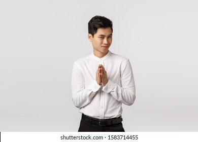 Mysterious and devious young good-looking asian businessman in white shirt, pants, rub hands and squinting sly, have secret evil plan, relish good deal, make money, standing white background