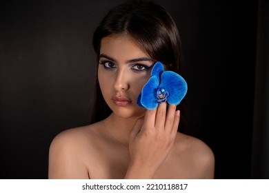 Mysterious Elegant, Romantic Brunette Naked Waist Up Woman With Make Up And Blue Orchid Flower Decoration Near Face Look At Camera On Black Studio Background. Perfume Flavor For Women. Skin, Body Care