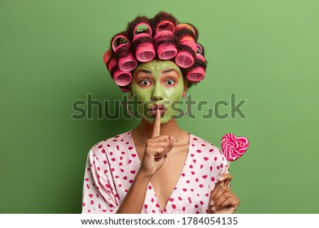 Mysterious ethnic woman wears green facial mask and hair rollers, makes silence gesture, tells secret, holds delicious heart shaped lollipop, makes hairstyle, wears dresssing gown, spends time at home