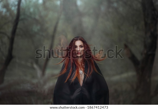 Mysterious fantasy gothic woman dark witch obsessed\
by evil. Red-haired Girl demon in black dress cape hood. Red hair\
flutters in wind. Dark dense deep forest background, trees. Scleral\
lenses on eyes