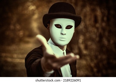 A mysterious gentleman in a white carnival mask and black tailcoat holds out his hand to the camera as an invitation, standing in an old autumn park. Mime, carnival. Halloween. Vintage style.