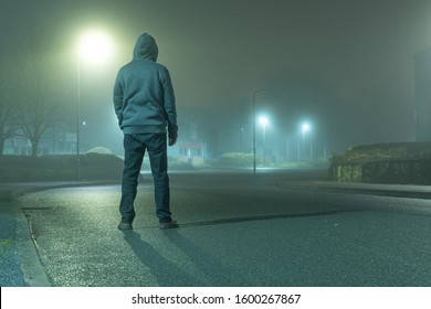 A Mysterious Hooded Figure With Back To Camera, Standing On A Road In A Light Industrial Urban Area. On A Moody, Foggy, Winters Night