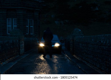 A mysterious hooded man, back to camera. Standing in front of car headlights. On a country lane at night