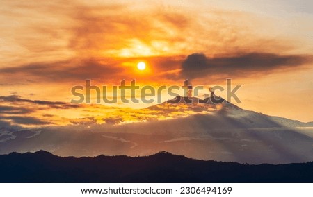 mysterious landscape of great erupting volcano with smoke from craters , clouds of gas and snow on slopes in orange light of sunset. eruption of vulcan
