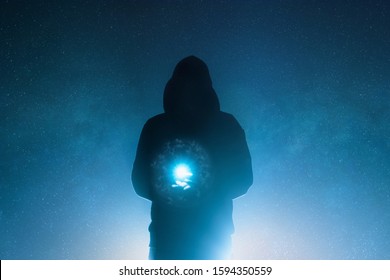 A mysterious moody hooded figure, holding a magical glowing light silhouetted against back light and a universe of stars.