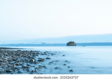 Mysterious old structure stands alone in calm waters at dusk near rocky shoreline under the serene evening sky - Powered by Shutterstock