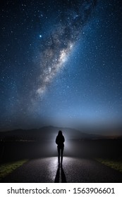 A mysterious person standing in the middle of the road looking into bright light with milky way starry night sky. 