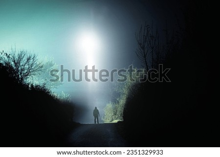 A mysterious scary UFO above a hooded figure. On a spooky country road. On a foggy winters night.