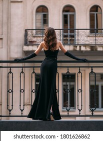 Mysterious silhouette of an elegant retro woman. A lady in long black evening dress stands on the balcony. Rear view, sexy back. Long vintage gloves.