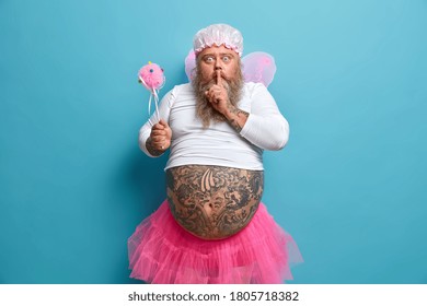 Mysterious Surprised Dad Plays Fairy On Birthday Party, Tells Secret, Makes Hush Gesture, Holds Magic Wand, Wears Pink Wings, Pleated Skirt, Shows Tattooed Belly, Isolated On Blue Wall. Home Party