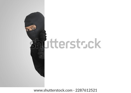 Mysterious thief wearing black hoodie hiding behind wall, sneaking, and looking for stolen goods. Crime concept. Isolated image on gray background