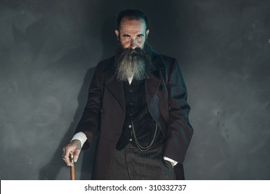 Mysterious vintage beard man in 1900 style fashion with cane against grey wall.