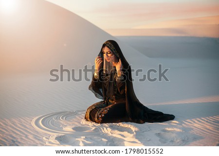 mysterious woman in abaya black long dress sits in desert nature. Luxury clothes, gold accessories hide face. Oriental beauty fashion model. Sand dunes background, orange sunset Art photo pretty lady