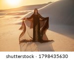 A mysterious woman in a black long dress stands in the desert. Luxurious clothes gold accessories hide the face. Oriental beauty fashion model. Sand dunes background orange sunset. hands raised to sky