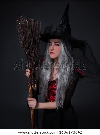 mysterious woman in black witch halloween costume and hat posing with broom over black background