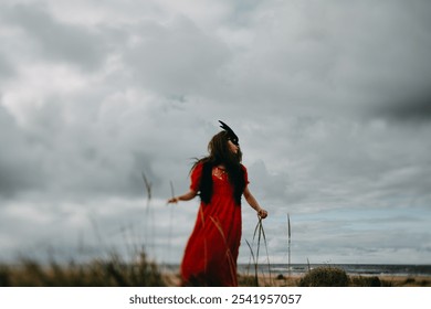 A mysterious woman in a red dress and dark feathered mask stands on a windy beach with an overcast sky. The moody setting and tall grasses create a surreal, enigmatic atmosphere - Powered by Shutterstock