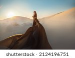 Mystery arabic woman in black long dress stands in desert long train silk fabric fly flutter in wind motion. clothes gold accessories hide face. Oriental fashion model. Sand dunes background sunset