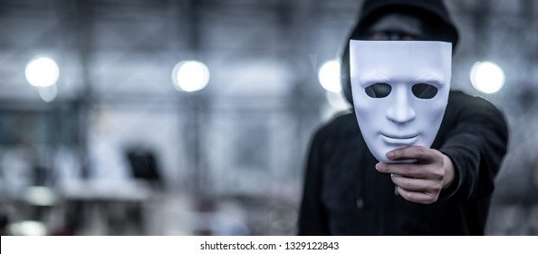 Mystery hoodie man with black mask holding white mask in his hand. Anonymous social masking or bipolar disorder concept.