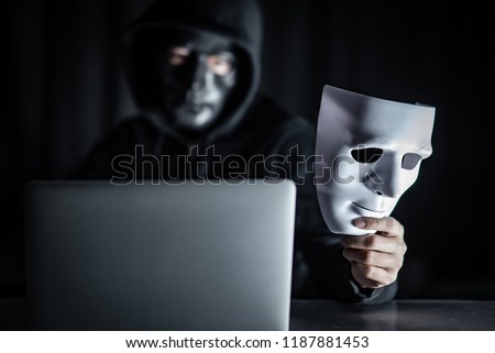 Mystery male hoodie hacker wearing black mask holding white mask with laptop computer on the table. Anonymous social masking. Ransomware cyber attack or internet security concepts