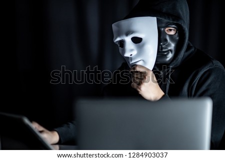 Mystery male hoodie hacker wearing black mask holding white mask sitting with laptop computer on the table. Anonymous social masking. Ransomware cyber attack or internet security concepts