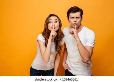 Mystery young couple posing together and showing silence gestures while looking at the camera over yellow background - Shutterstock ID 1024688614