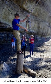 Mystic Beach, Vancouver Island, British Columbia, September 11, 2016: A Man In A Blue Shirt Stands On A Log As He Gets Ready To Jump Onto A Rope Swing On The Beach, While Others Watch. 