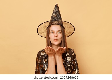 Mystic and magical holiday. Black witch theme party. Romantic young girl wearing black halloween garment and cone posing isolated over beige background sending air kissing over plams flirting