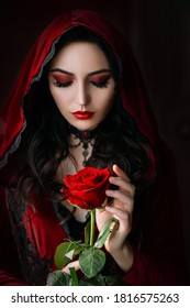 Mystical Beautiful Woman In A Gothic Costume Of A Medieval Vampire In A Hood. An Adult Girl Holds A Rose Flower In Her Hands. Beautiful Face Red Makeup, Attractive Lips. Halloween Party Image