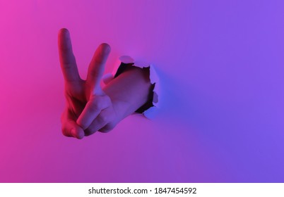 Mystical halloween concept  Hand shows V gesture through torn hole Creative pop art pink blue neon color  Trendy gradient illumination  Night light