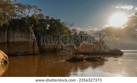Mystical morning landscape on the river. Limestone rocks are reflected in the red-brown water. The sun shines in the sky through the clouds.  Haze in the distance. Madagascar. Manambolo River.