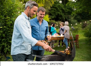 n a summer evening,  two men  in their forties prepares a barbecue for  friends gathered around a table in the garden