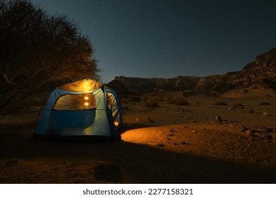 Nahal Menagen, Big Dune, Timna Park, Israel Camping at Nahal Menagan in blue tent glowing from yellow lamps no a full moon night in the desert with mountains and acacia tree - Powered by Shutterstock